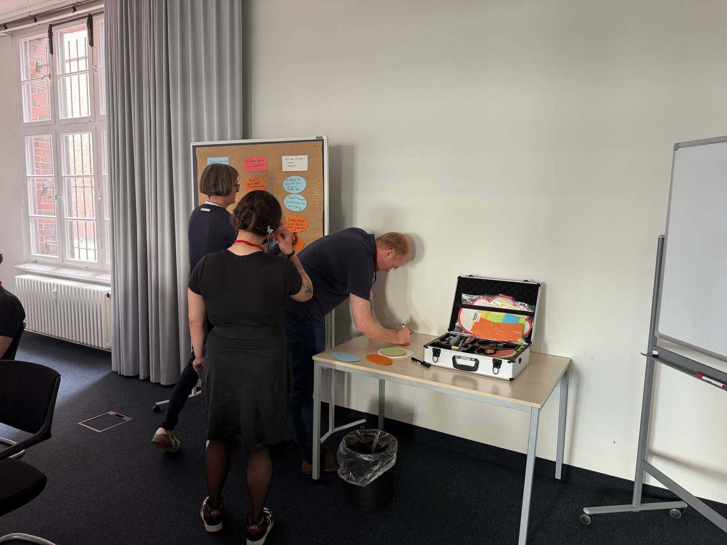 3 Personen stehen vor einem Tisch mit einem aufgeklappten Koffer. In dem Koffer befinden sich Büromaterialien.