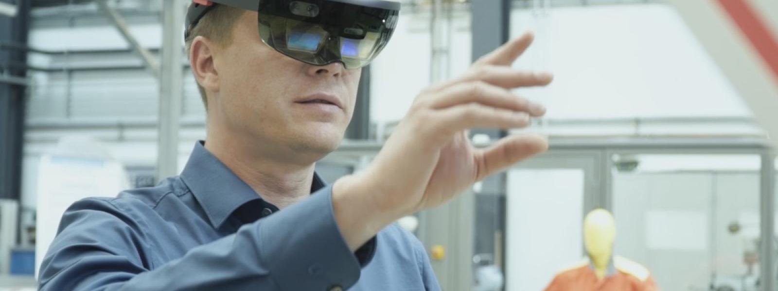 Eine Person mit aufgesetzter Augmented Reality Brille steht in einer Industriehalle und hält einen Arm ausgestreckt vor dem Körper