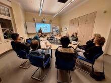 Raumansicht mit Konferenztisch um den Menschen verteilt sitzten und ein Vortragender vor Kopf